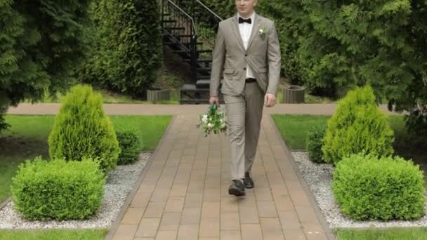 Groom desce o beco entre arbustos com um buquê de casamento para sua noiva amada — Vídeo de Stock