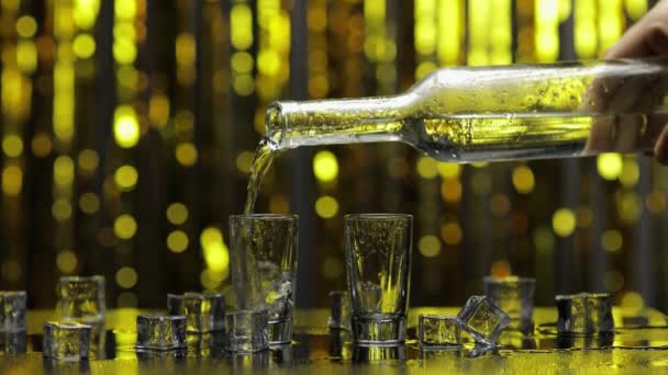 Bartendern häller fryst vodka från flaskan i glas. Is kuber mot glänsande guld parti bakgrund — Stockvideo