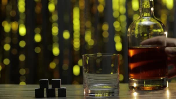 Nalití zlaté whisky, koňaku nebo brandy z láhve do skla s ledovými kameny na stole — Stock video