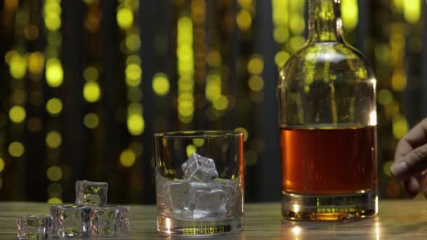 Pouring of golden whiskey, cognac or brandy from bottle into glass with ice cubes. Shiny background — Stock Video