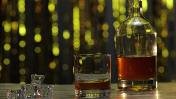 Barman dodaje kostki lodu do szklanki ze złotą whisky, koniakiem lub brandy na stole. Błyszczące tło — Wideo stockowe