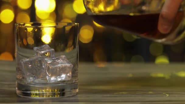 Arany whisky, konyak vagy brandy üvegből üvegbe öntése jégkockákkal. Fényes háttér — Stock videók