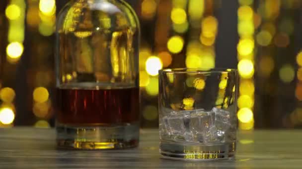 Menuangkan wiski emas, cognac atau brendi dari botol ke kaca dengan es batu. Latar belakang berkilau — Stok Video