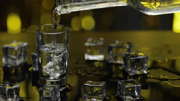 Barman vierta el vodka congelado de la botella en un vaso de chupito. Cubos de hielo contra fondo brillante fiesta de oro — Foto de Stock