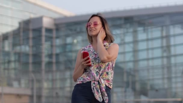 Mujer turista alegre en gafas de sol rosas bailando y escuchando música en auriculares. Moviéndose en danza — Vídeo de stock