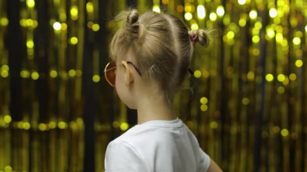 Kinderspel duimen omhoog, glimlachend, kijkend naar de camera. meisje poseren op de achtergrond met folie gouden gordijn — Stockvideo
