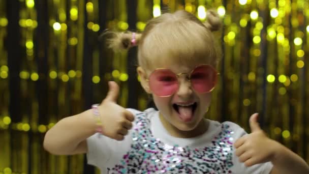 Kinderspel duimen omhoog, glimlachend, kijkend naar de camera. meisje poseren op de achtergrond met folie gouden gordijn — Stockvideo