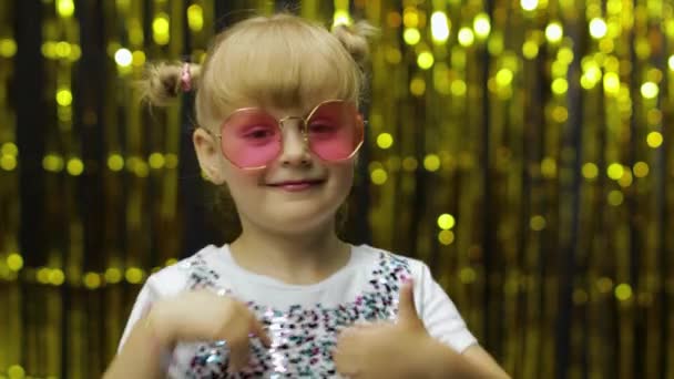 Child show thumbs up. Smiling, looking at camera. Girl posing on background with foil golden curtain — Stock Video