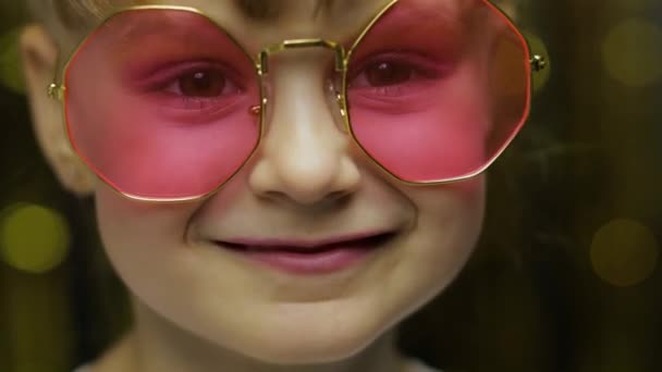 Κοντινό πρόσωπο του παιδιού. Χαμογελώντας, κοιτάζοντας την κάμερα. Κορίτσι με ροζ γυαλιά ηλίου που ποζάρει — Αρχείο Βίντεο