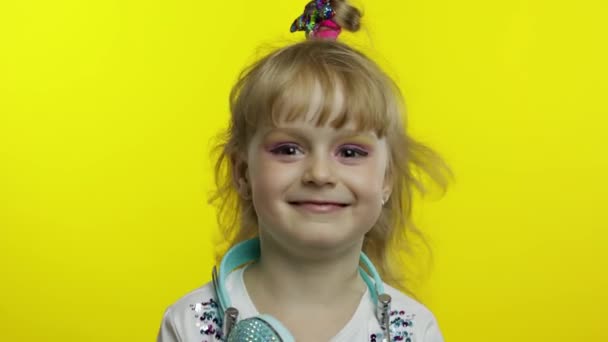 Lachend kind, kijkend naar de camera. Meisje met hoofdtelefoon op de nek poseren op gele achtergrond — Stockvideo