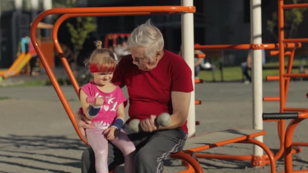 Kleindochter en grootvader doen fitnessoefeningen met halters. Senior man met kind kind meisje — Stockvideo