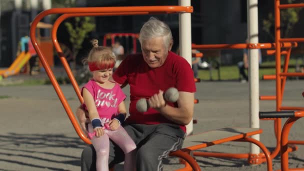 Kleindochter en grootvader doen fitnessoefeningen met halters. Senior man met kind kind meisje — Stockvideo