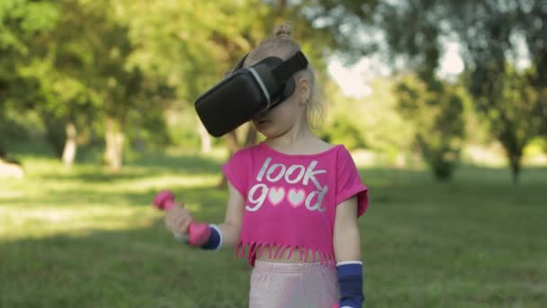 Athletic child girl in VR headset helmet making fitness workout exercises with dumbbells in park — Stock Video