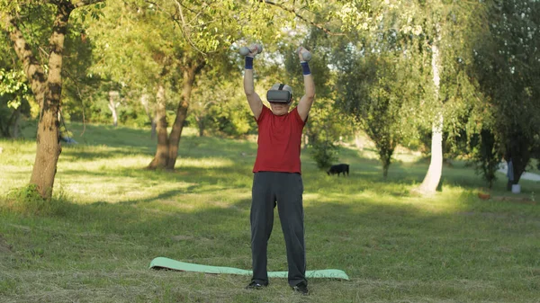 Sénior velho avô homem em VR capacete de fone de ouvido fazendo exercícios de fitness com halteres no parque — Fotografia de Stock