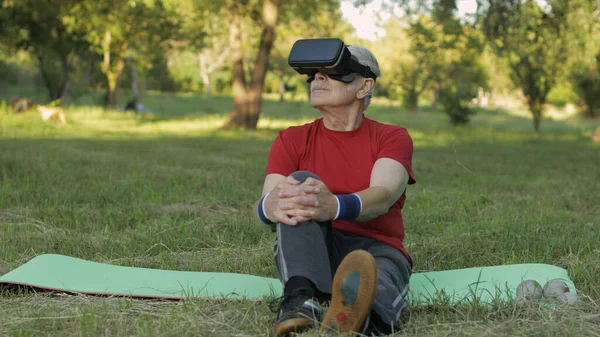 Elderly man in VR headset helmet sitting on sport mat. Online sport workout for senior people