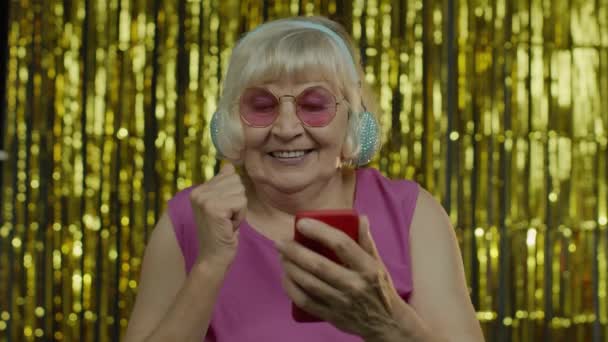 Стильна щаслива старша жінка тримає мобільний телефон, слухає музику в навушниках і танцює — стокове відео