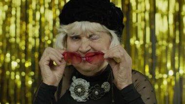 Sevimli yaşlı kadın kırmızı güneş gözlüklerine dokunuyor ve neşeyle göz kırpıyor, flört ediyor ve göz kırpıyor.