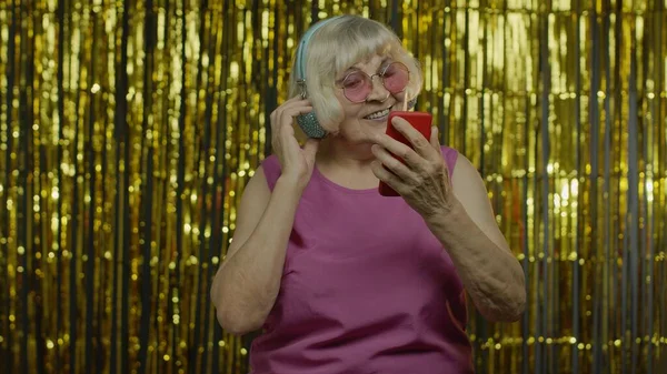 Elegante anciana feliz sosteniendo el teléfono celular, escuchando música en los auriculares y bailando —  Fotos de Stock