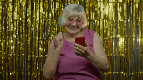 Elegante anciana feliz sosteniendo el teléfono celular, escuchando música en los auriculares y bailando —  Fotos de Stock