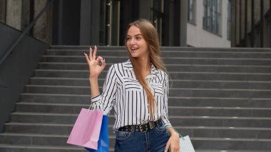 Alışveriş torbaları tutan şık bir kız ve tamam işareti gösteriyor. İyi indirimli tatil indirimleriyle neşe içinde