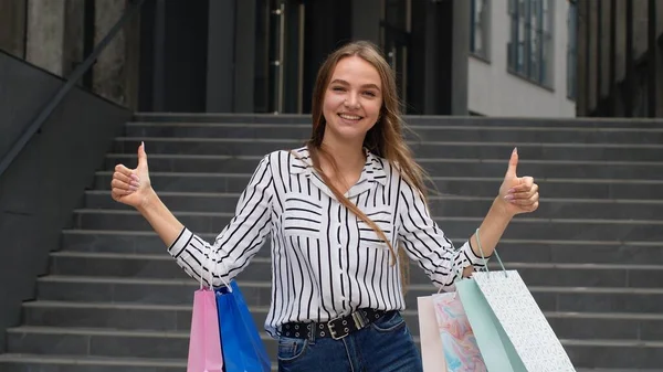 Alışveriş torbaları tutan ve baş parmak gösteren modaya uygun bir kız. İyi indirimli tatil indirimleriyle neşe içinde