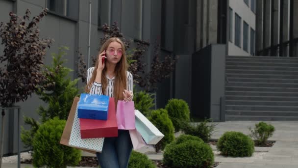 Gadis muda positif dengan tas berbicara di telepon genggam tentang penjualan di pusat perbelanjaan di Black Friday — Stok Video