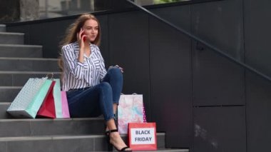 Merdivenlerde oturan ve cep telefonuyla Kara Cuma 'daki alışveriş merkezinde indirimden bahseden bir kız.