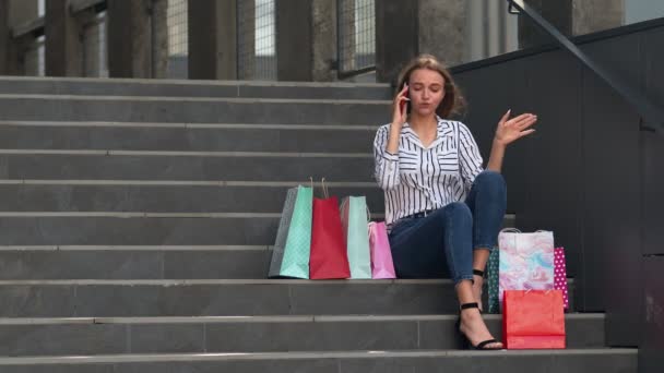 Dziewczyna siedzi na schodach z torbami rozmawiając przez telefon komórkowy o sprzedaży w centrum handlowym w Czarny Piątek — Wideo stockowe