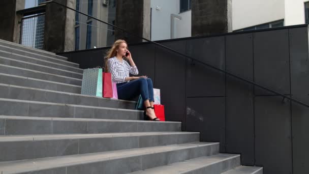 Chica sentada en las escaleras con bolsas hablando en el teléfono móvil sobre la venta en el centro comercial en el Viernes Negro — Vídeos de Stock