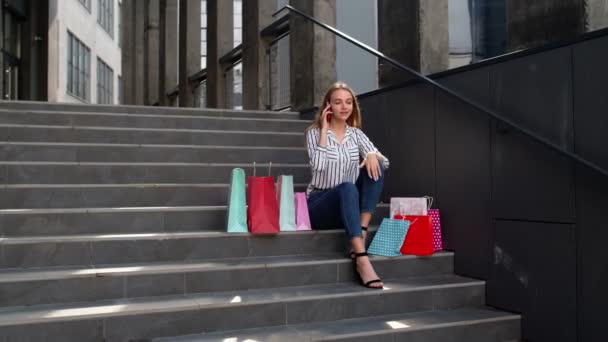 Fille assise sur les escaliers avec des sacs parler sur téléphone mobile à propos de la vente dans le centre commercial Black Friday — Video