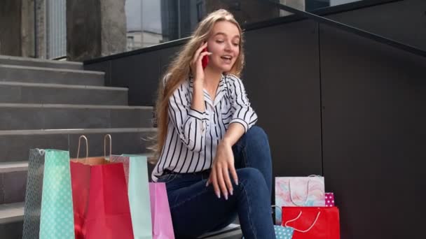 Chica sentada en las escaleras con bolsas hablando en el teléfono móvil sobre la venta en el centro comercial en el Viernes Negro — Vídeos de Stock