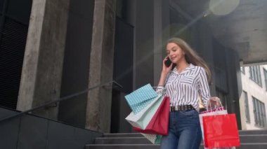 Alışveriş merkezinden alışveriş torbalarıyla gelen ve cep telefonuyla alışverişler hakkında konuşan bir kız.
