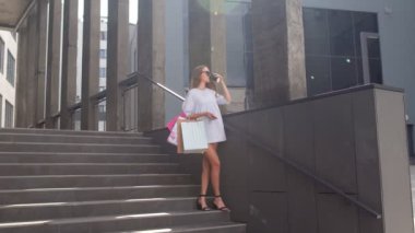 Telefonu olan kız alışveriş merkezinin merdivenlerinde renkli alışveriş torbalarıyla oturuyor ve kahve içiyor.