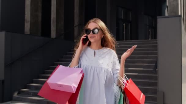 ショッピングバッグを持ってモールから歩き、ブラック・フライデーでの販売について携帯電話で話す女の子 — ストック動画
