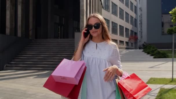 Chica caminando desde el centro comercial con bolsas de compras y hablando por teléfono móvil sobre las compras — Vídeos de Stock