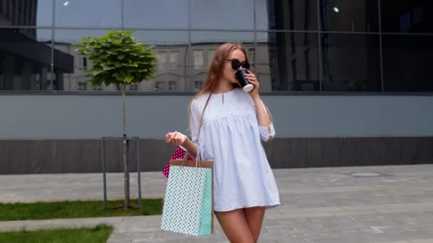 Menina ficar perto do shopping com sacos de compras e beber café. Relaxante depois de fazer compras na loja de moda — Vídeo de Stock