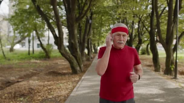 Άντρας ηλικιωμένος τρέχει κατά μήκος του δρόμου στο πάρκο. Ώριμος δρομέας προπόνηση, μουσική ακρόασης — Αρχείο Βίντεο