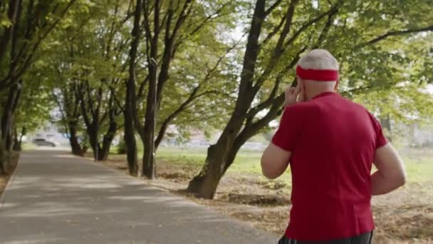 Bakifrån av äldre gubbe kör, träna cardio i parken och lyssna musik på hörlurar — Stockvideo