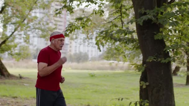 Старший старик бегает, тренирует кардио в парке и использует Bluetooth наушники, слушает музыку — стоковое видео