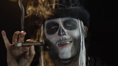 Şapkalı, iskelet makyajlı ürkütücü bir adam. Puro içen, surat yapan, kameraya bakan bir adam.