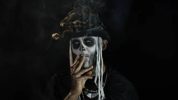 Sinister man with horrible Halloween skeleton makeup smoking cigar, making faces