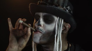 Şapkalı, iskelet makyajlı ürkütücü bir adam. Puro içen, surat yapan, kameraya bakan bir adam.