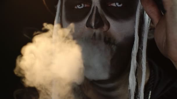 Primer plano del hombre siniestro con el maquillaje del cráneo haciendo caras y exhalando humo de cigarrillo de su boca — Vídeos de Stock