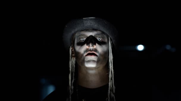 Złowieszczy człowiek ze szkieletowym makijażem, wyłaniający się z ciemności, gdy pada na niego światło, robiąc twarze. — Wideo stockowe