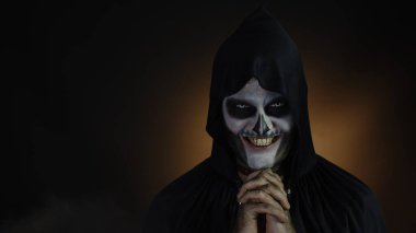 Kapüşonlu iskelet makyajlı ürkünç, deli bir adam kızgın suratlar yapıyor, başını sallıyor, korkutmaya çalışıyor.