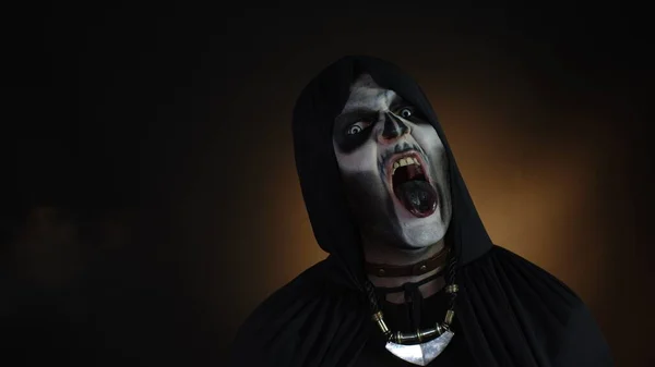 Gruseliger Mann mit Skelett-Make-up in Kapuze. Guy Henker macht wütende Gesichter, schaut in die Kamera und versucht zu erschrecken. Voodoo-Rituale. Baron Samstag. Themenparty zu Halloween. Schwarzer Hintergrund — Stockbild Gruseliger Mann mit Skelett-Make-up in Kapuze macht wütende Gesichter, blickt in die Kamera, versucht Angst zu bekommen — Stockfoto