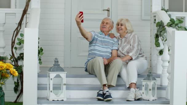 Старші люди похилого віку, які користуються мобільним телефоном для відеодзвінків, роблять селфі вдома на ґанку. — стокове відео