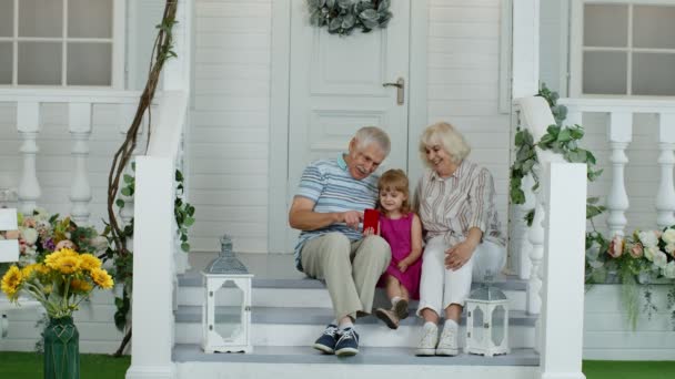 Äldres par sitter hemma med barnbarn på verandan. Använda mobiltelefon för videosamtal online — Stockvideo