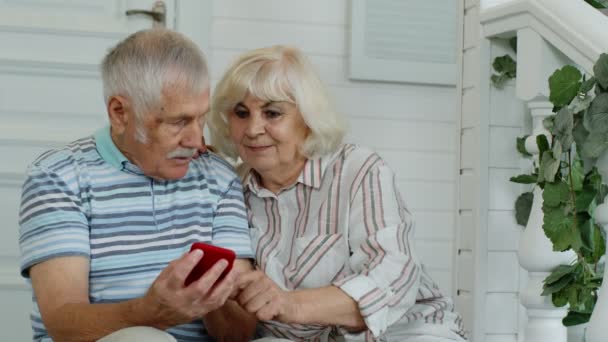 Старша подружня пара кавказьких старців користується мобільним телефоном вдома. Насолоджуйся онлайн-магазинами, новинами — стокове відео