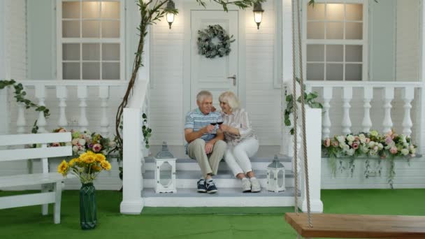 Satisfecho senior ancianos caucásico pareja sentado y beber vino en porche escaleras en casa — Vídeos de Stock